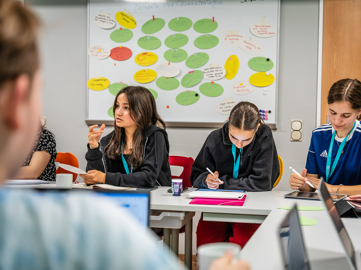 Einige Jugendliche sitzen an Tischen und arbeiten. Im Hintergrund ist ein Whiteboard, an dem bunte Zettel befestigt sind, zu sehen.