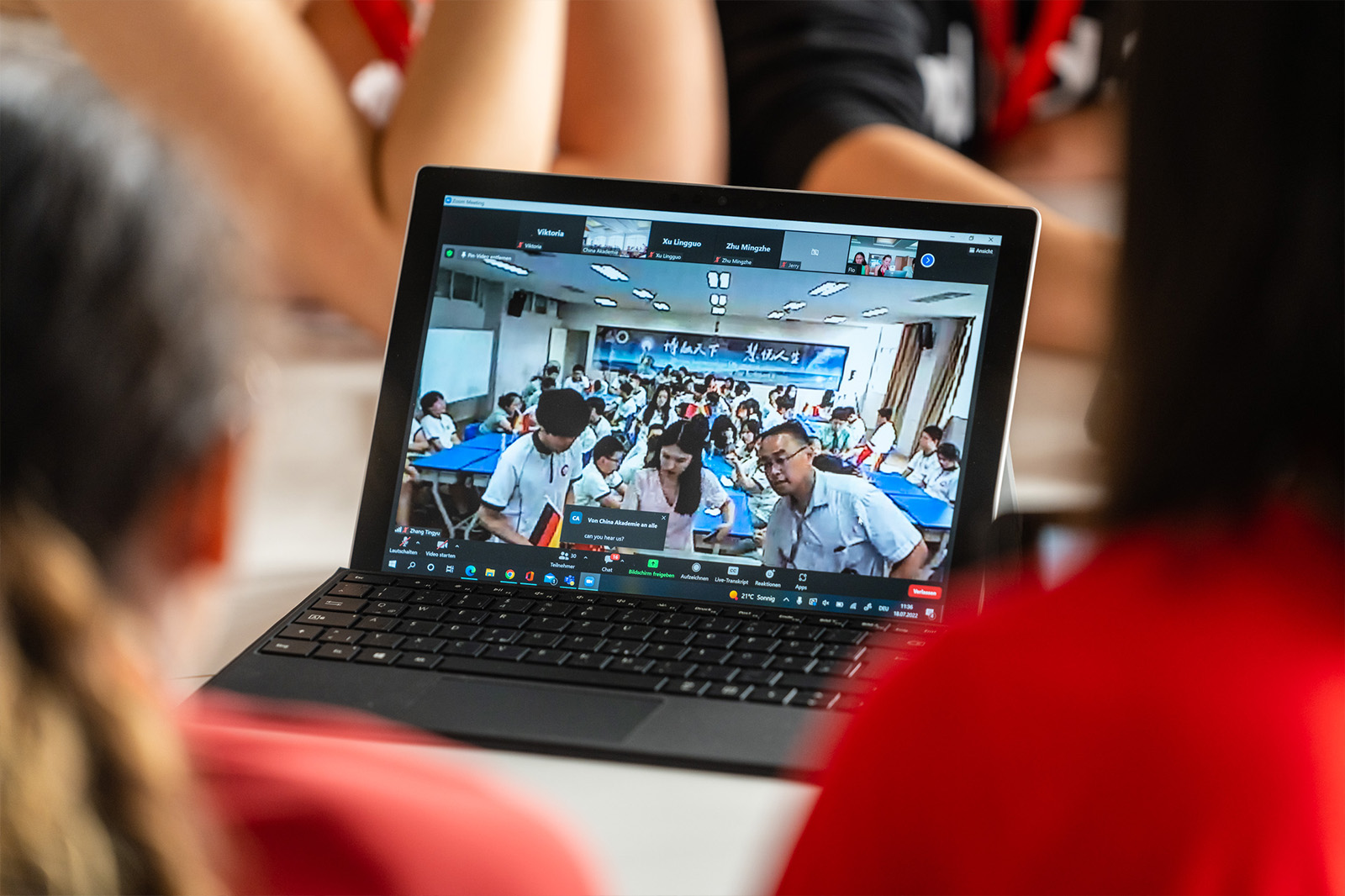 Zwei Jugendliche sitzen vor einem aufgeklappten Laptop, auf dem man eine Gruppe chinesischer Schülerinnen und Schüler erkennt.