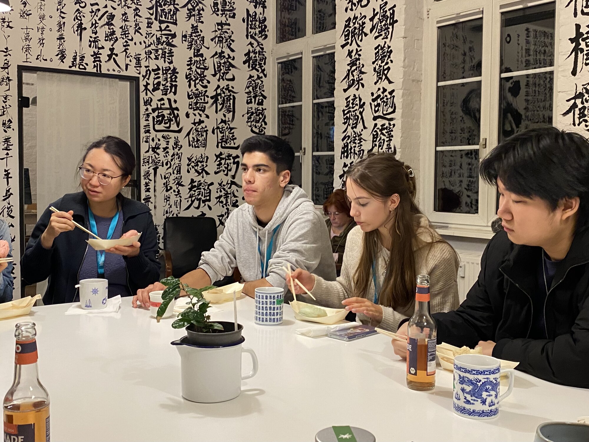 Deutsche und chinesische Schüler:innen sitzen gemeinsam an einem Tisch und essen Baozi. Die Wand im Hintergrund ist vollständig mit chinesischer Kalligrafie bemalt.