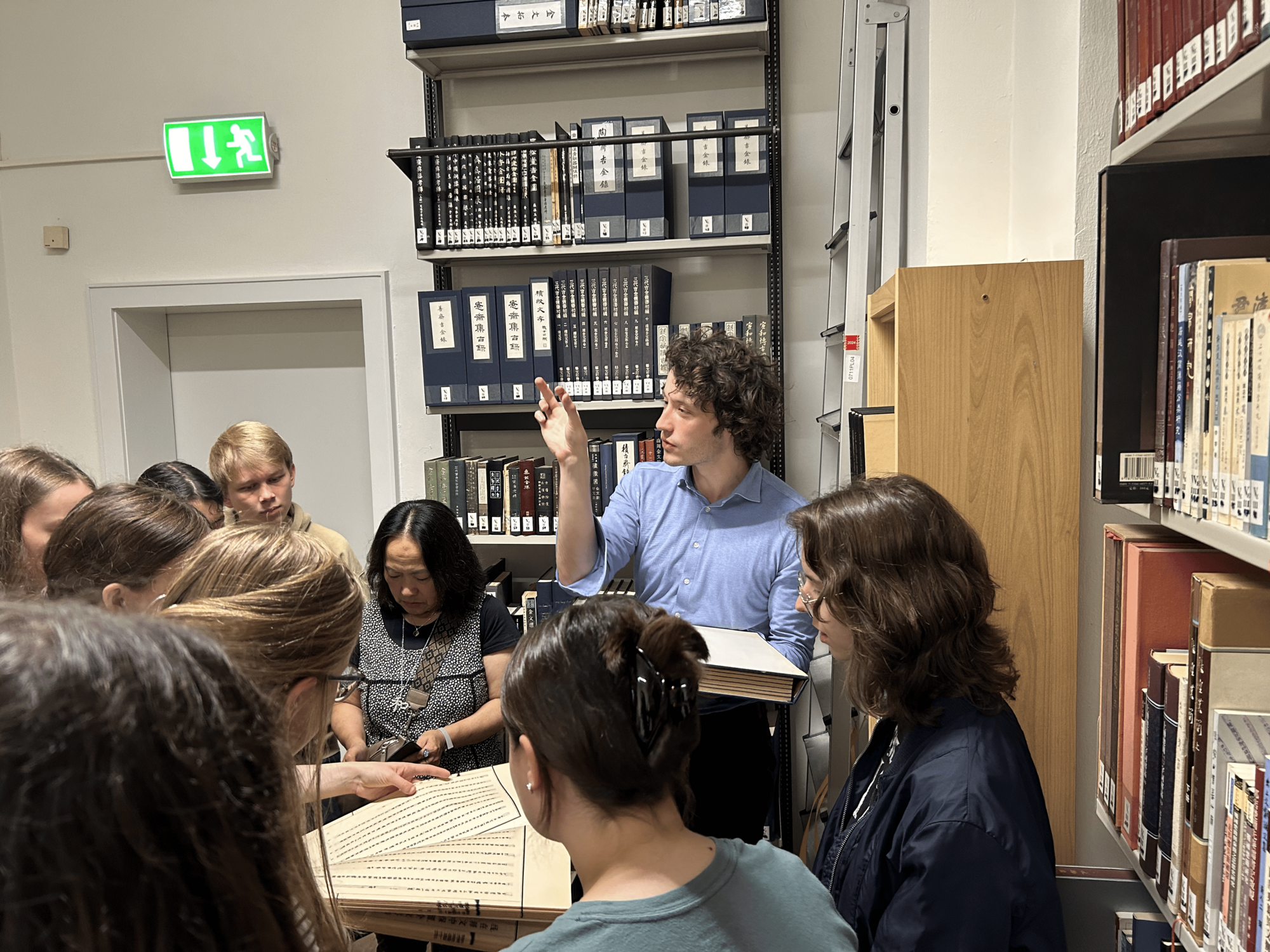 Jugendliche und ein junger Mann stehen in einer Bibliothek und schauen sich ein Buch mit chinesischen Schriftzeichen an. Im Hintergrund sind gefüllte Bücherregale.