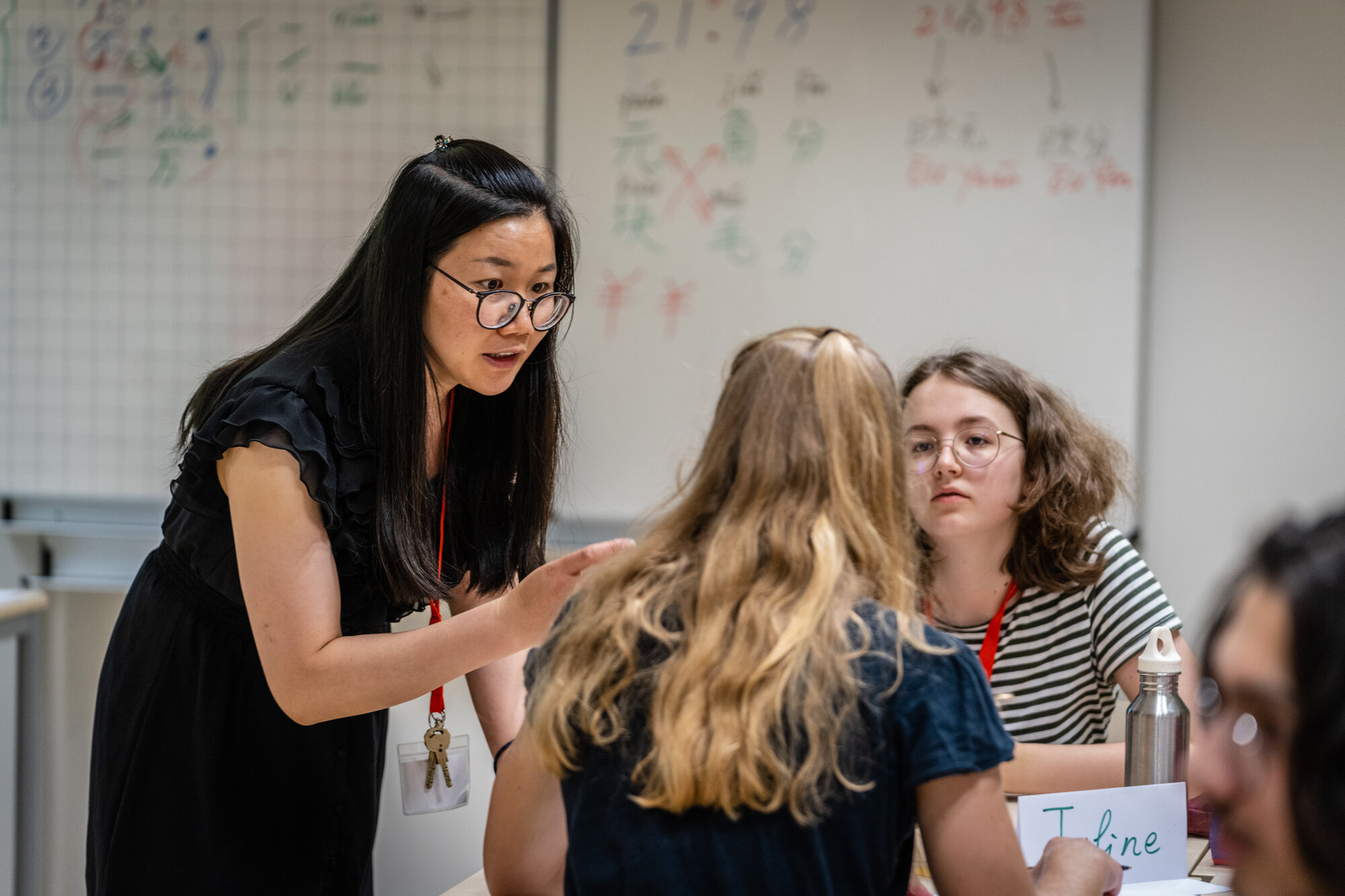 Eine Person mit asiatischen Gesichtszügen steht an einem Tisch mit jungen Schüler:innen. Im Hintergrund ist ein Whiteboard mit chinesischen Schriftzeichen zu sehen.