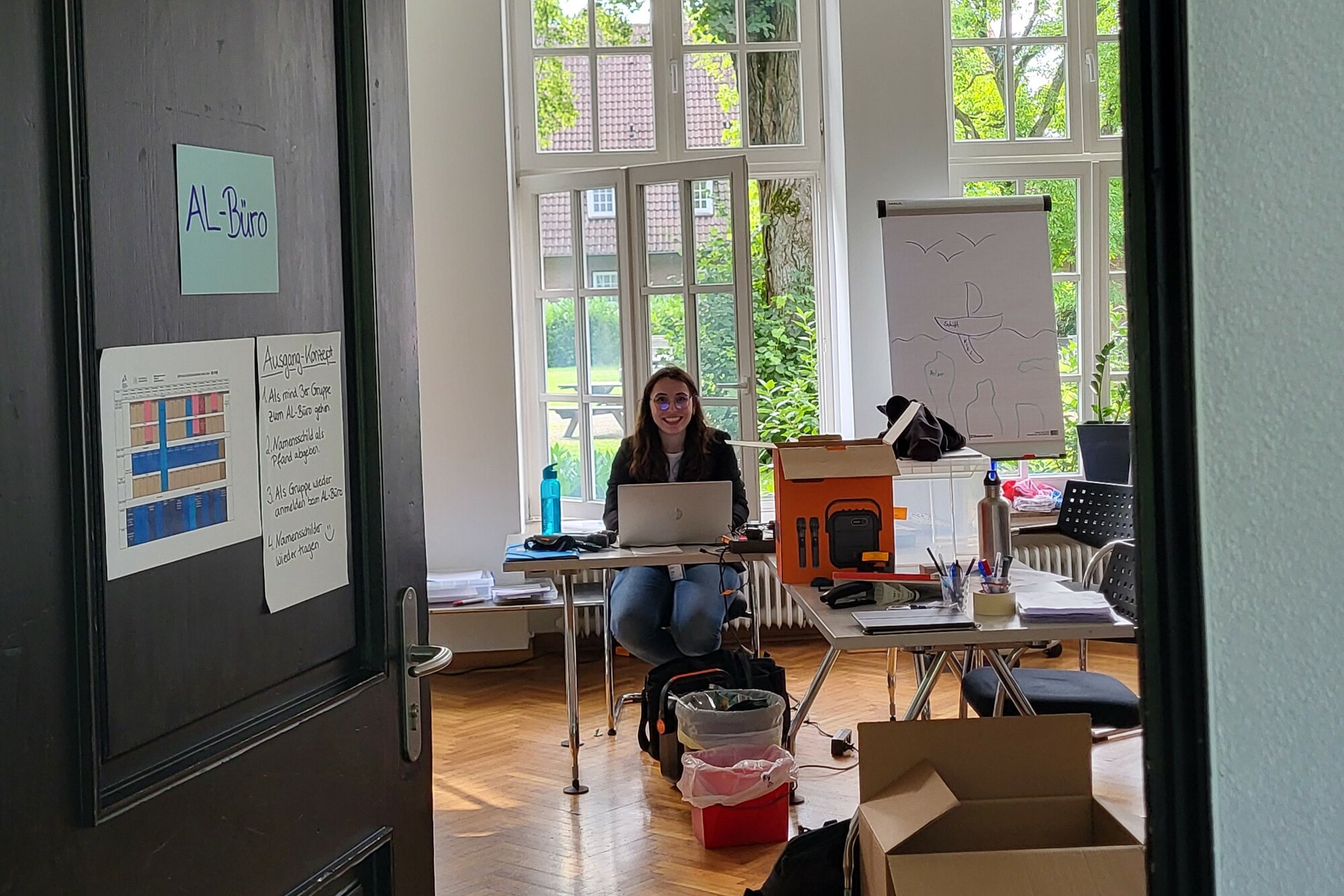 In einem unordentlichen Büroraum mit großen Fenstern sitzt eine Frau an einem Laptop.