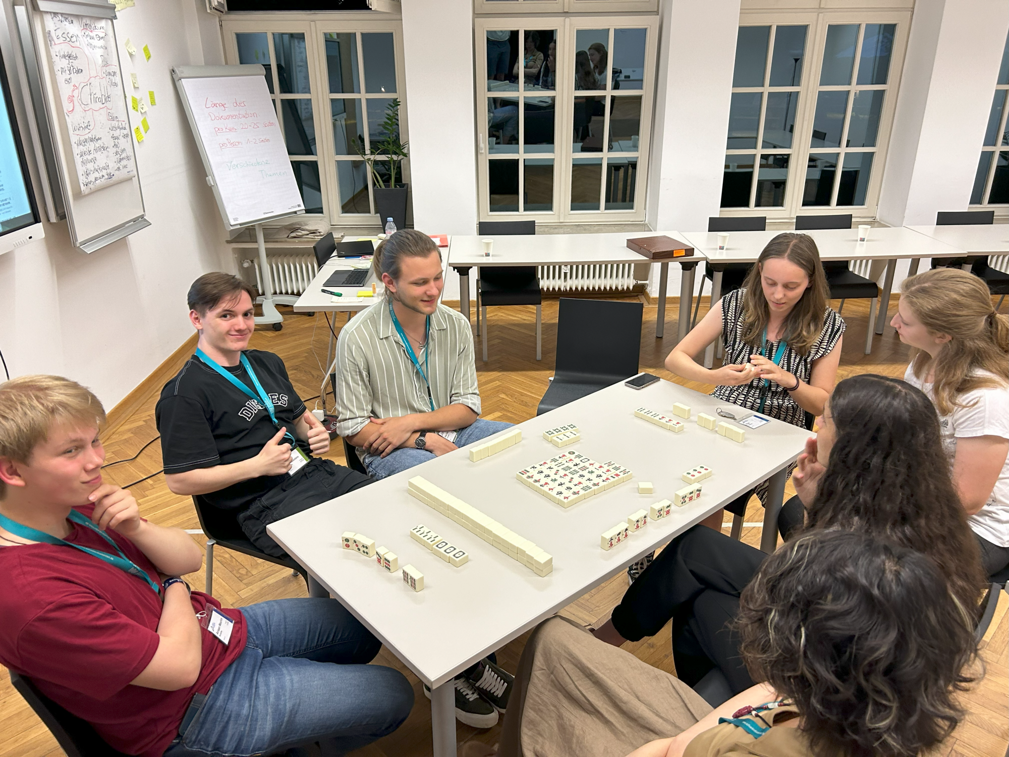Sechs Jugendliche sitzen an einem Tisch und spielen Mahjong. Im Hintergrund sind ein Flipchart, ein Laptop, weitere Tische und große Fenster zu sehen.