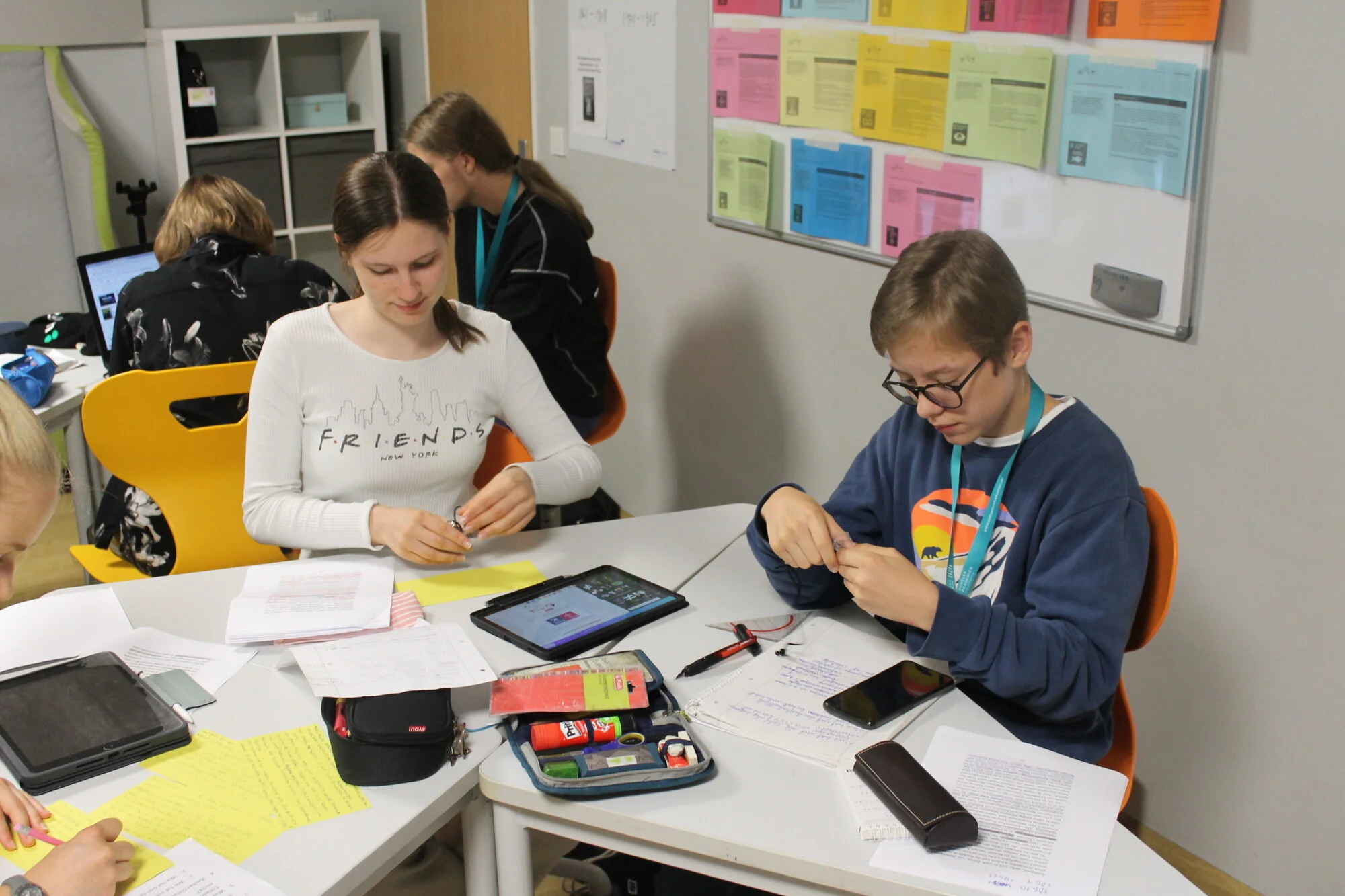 Im Vordergrund sitzen Schüler:innen an Tischen mit Notizblöcken, Tablets und Schreibmaterial. Im Hintergrund hängt ein buntes Plakat an der Wand.