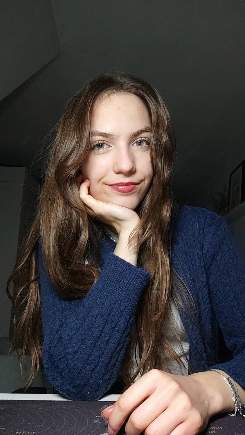 Porträt einer jugendlichen Frau mit langen braunen Haaren und grünen Augen. Sie stützt den Kopf auf eine Hand und lächelt leicht in die Kamera.