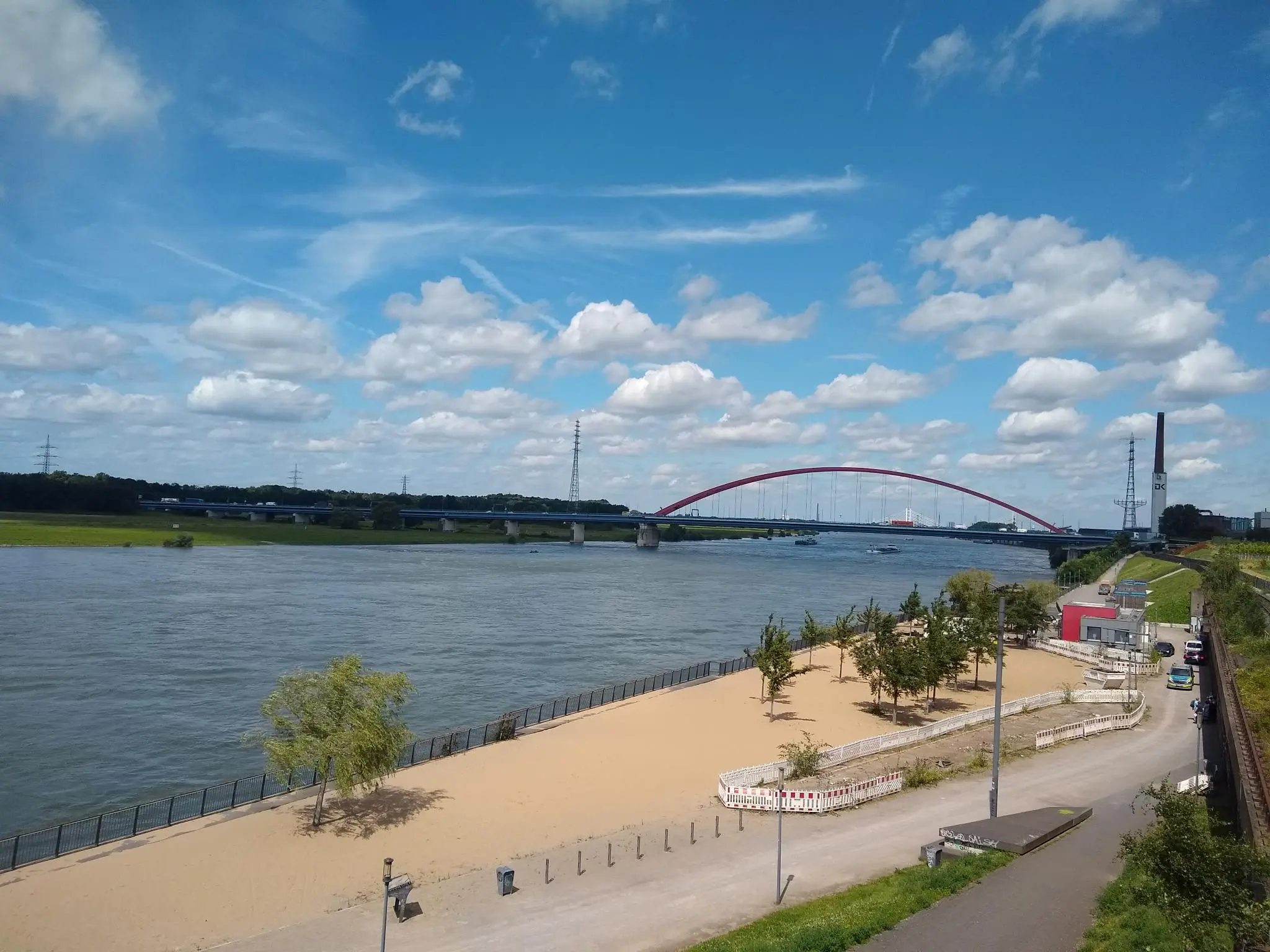 Aus der Vogelperspektive ist ein sandiges Flussufer mit einzelnen Bäumen zu sehen. Im Hintergrund sind ein Fluss und eine große Brücke zu erkennen.