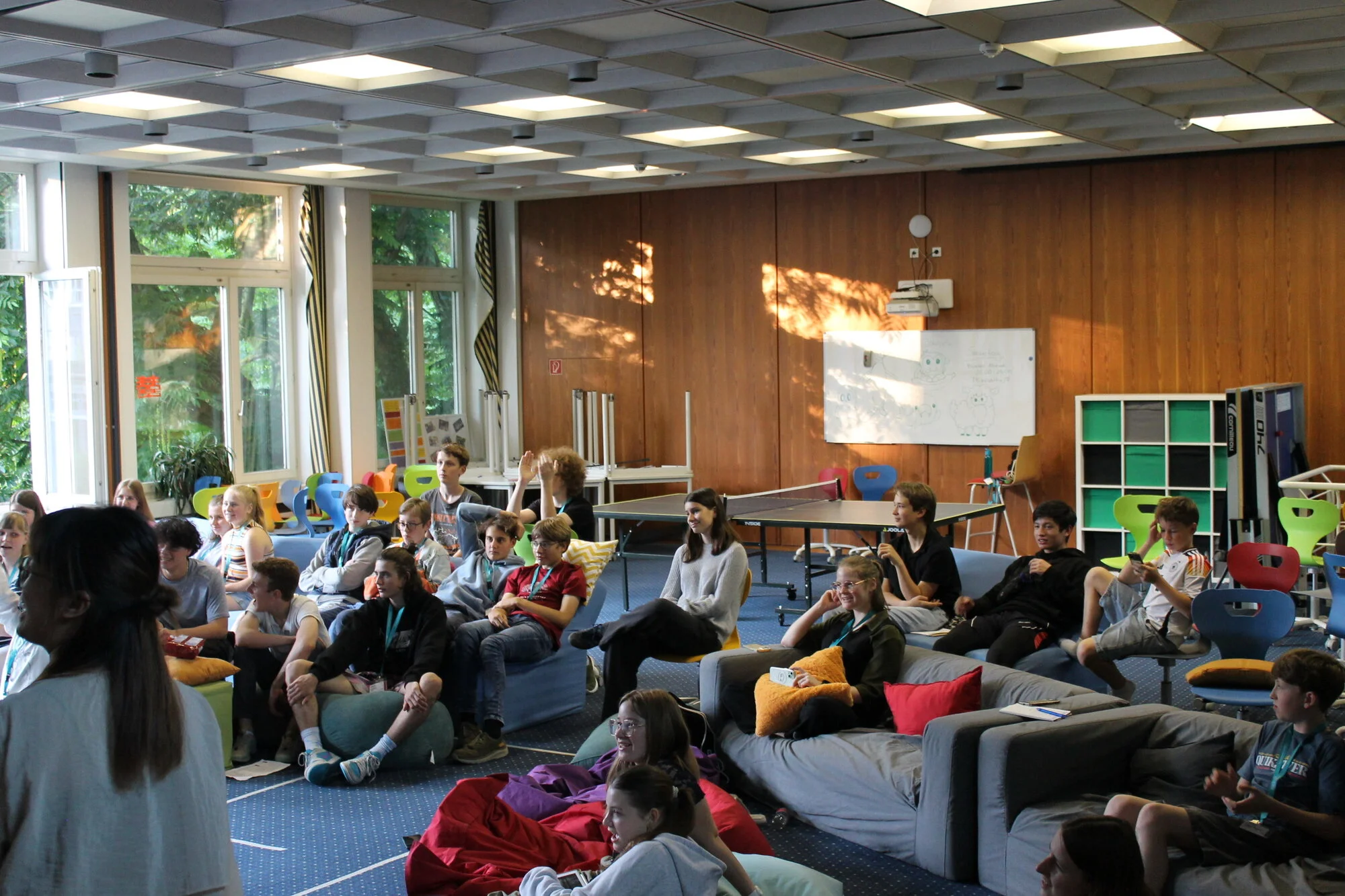 In einem hellen, großen Raum sitzen viele Jugendliche auf bequemen Sitzgelegenheiten wie Sofas und Sitzsäcken.