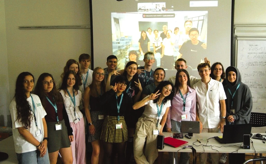 Eine Gruppe Jugendliche posiert für ein Foto. Im Hintergrund ist ein Bild einer Schulgruppe an die Wand gebeamt, die ebenfalls posiert.