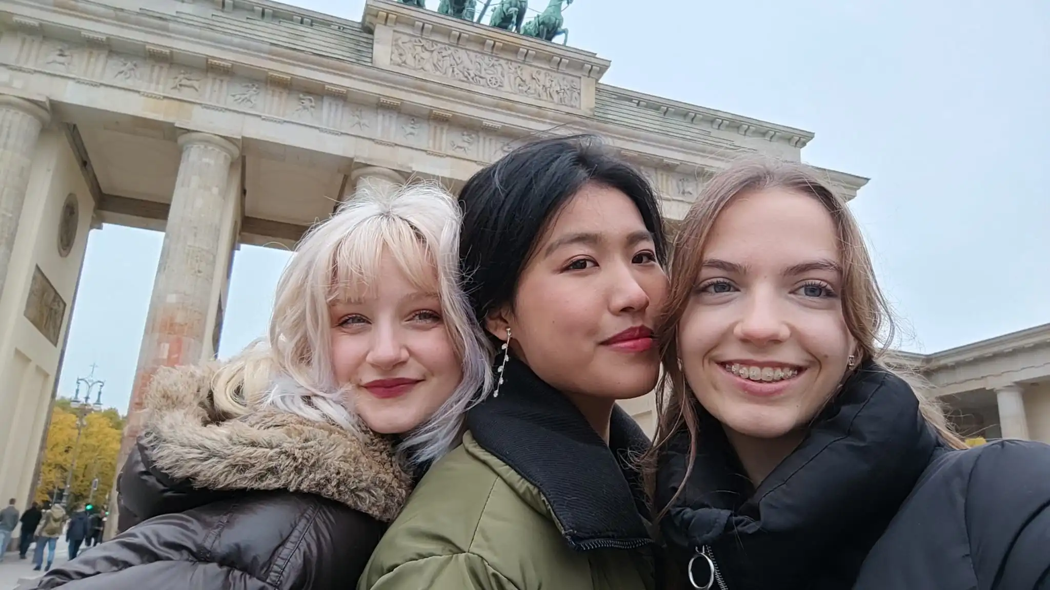 Drei jugendliche Frauen in winterlicher Kleidung posieren für ein Selfie und lächeln in die Kamera. Im Hintergrund steht das Brandenburger Tor.
