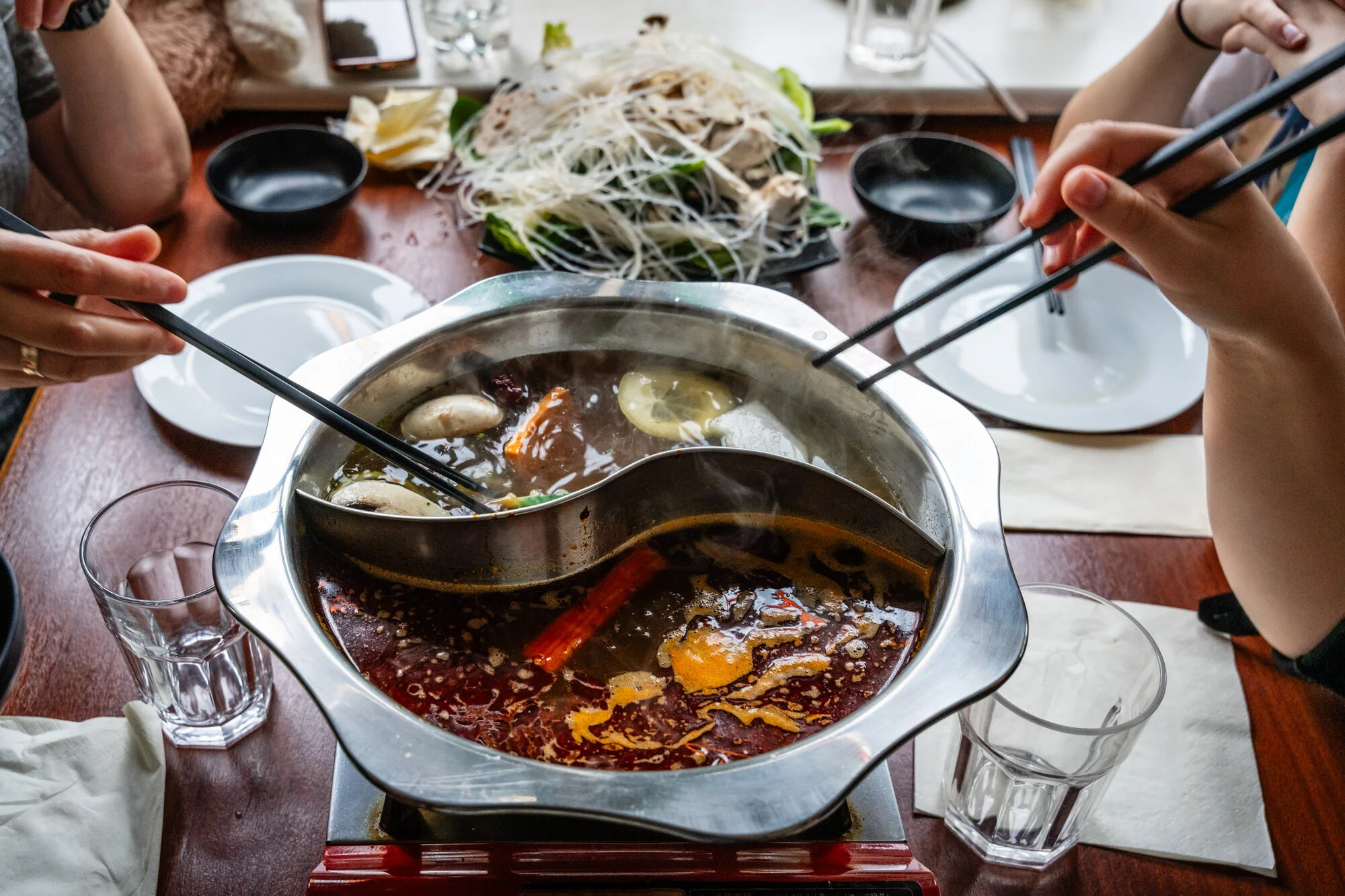Auf einem Tisch steht ein Hotpot mit dampfender Brühe. Darum herum stehen Gläser, Schalen und Teller mit Zutaten. Zwei Hände greifen mit Stäbchen in den Topf, um Speisen herauszuholen.