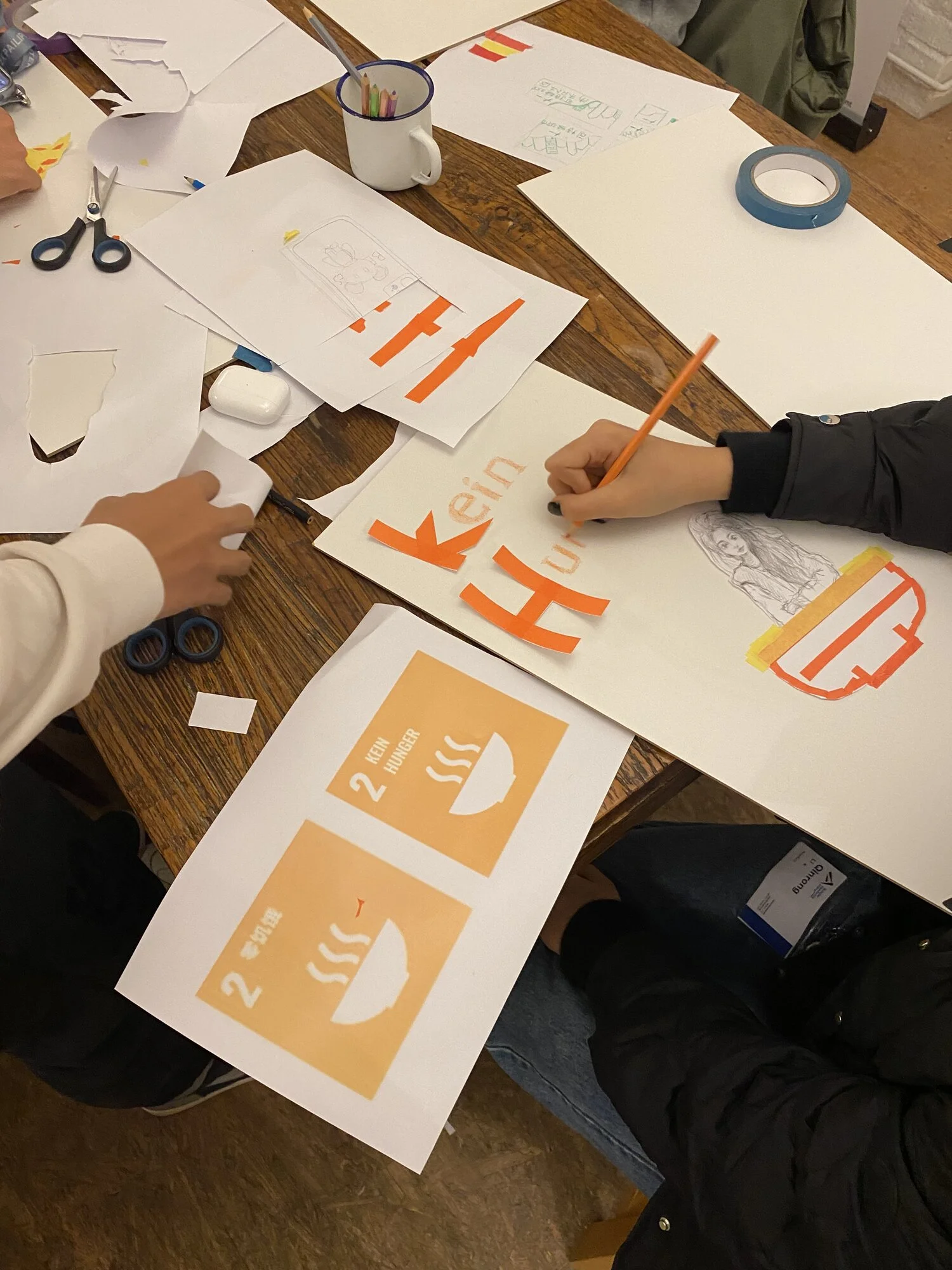 Auf einem Tisch liegen Papier, Abbildungen der SDGs, eine Schere und ein Klebestift. Mehrere Hände halten Stifte und basteln.