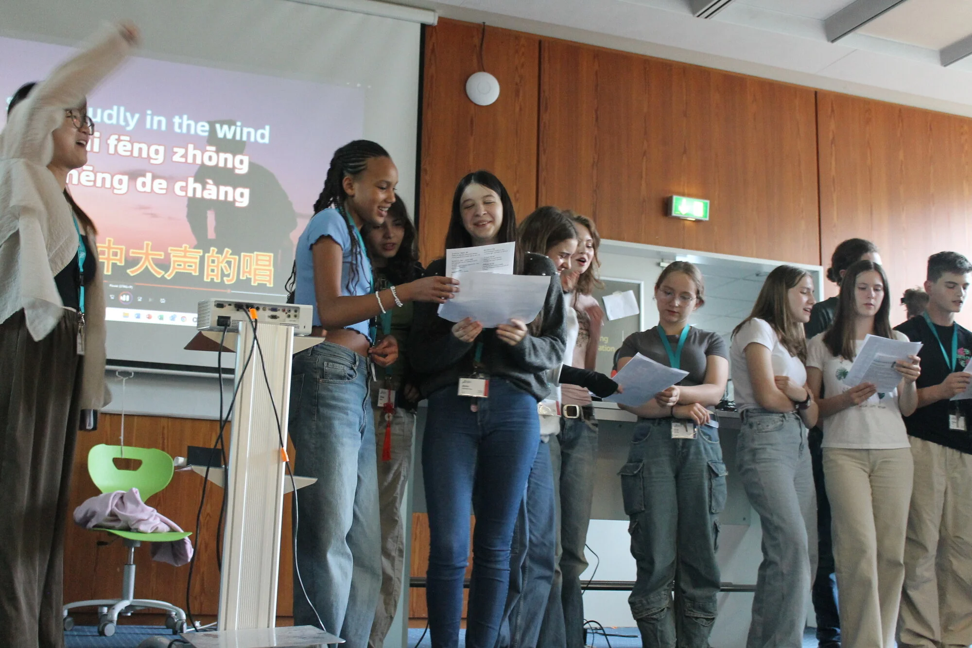 Mehrere Jugendliche stehen nebeneinander vor einer Wand. Sie halten Liedtexte in den Händen und singen. An die Wand hinter ihnen wird per Beamer der Text einer Karaoke-Version projiziert. Am Bildrand steht eine Frau und gibt mit den Händen Dirigierzeichen.