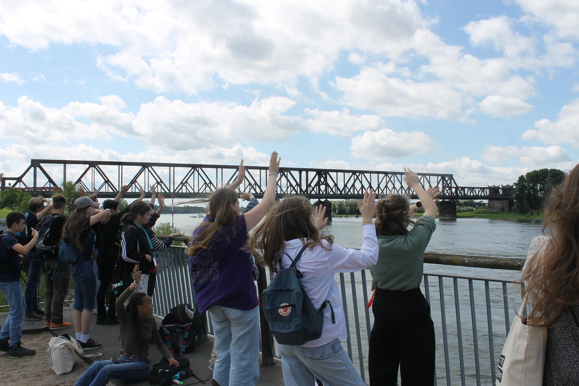 Junge Menschen stehen an einem Geländer, schauen auf den Fluss und winken. Im Hintergrund sind eine große Brücke und der Fluss zu sehen.