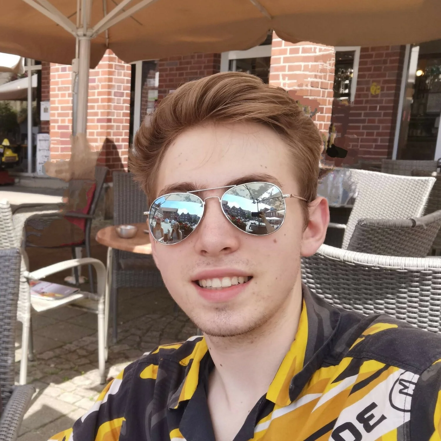 Porträt eines jugendlichen Mannes mit braunen Haaren. Er trägt eine große, spiegelnde Sonnenbrille und ein orange-schwarzes Hemd. Im Hintergrund stehen Stühle und Sonnenschirme.