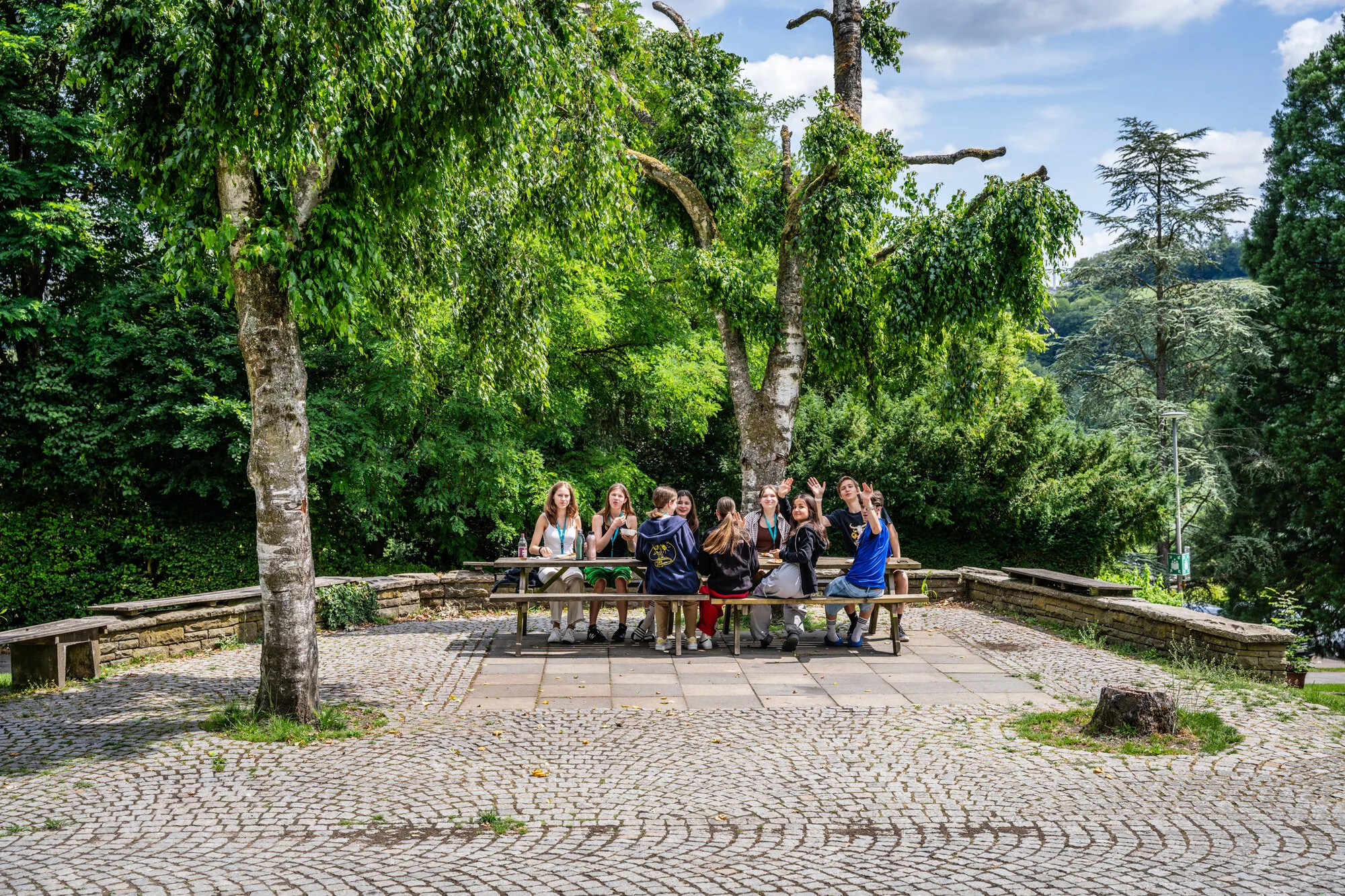 Jugendliche sitzen an einem Holztisch auf einem gepflasterten Platz. Im Hintergrund stehen grüne, dicht belaubte Bäume.