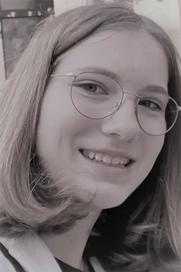 Ein jugendliches Mädchen mit Brille und schulterlangem Haar lächelt in die Kamera. Das Bild ist schwarz-weiß.